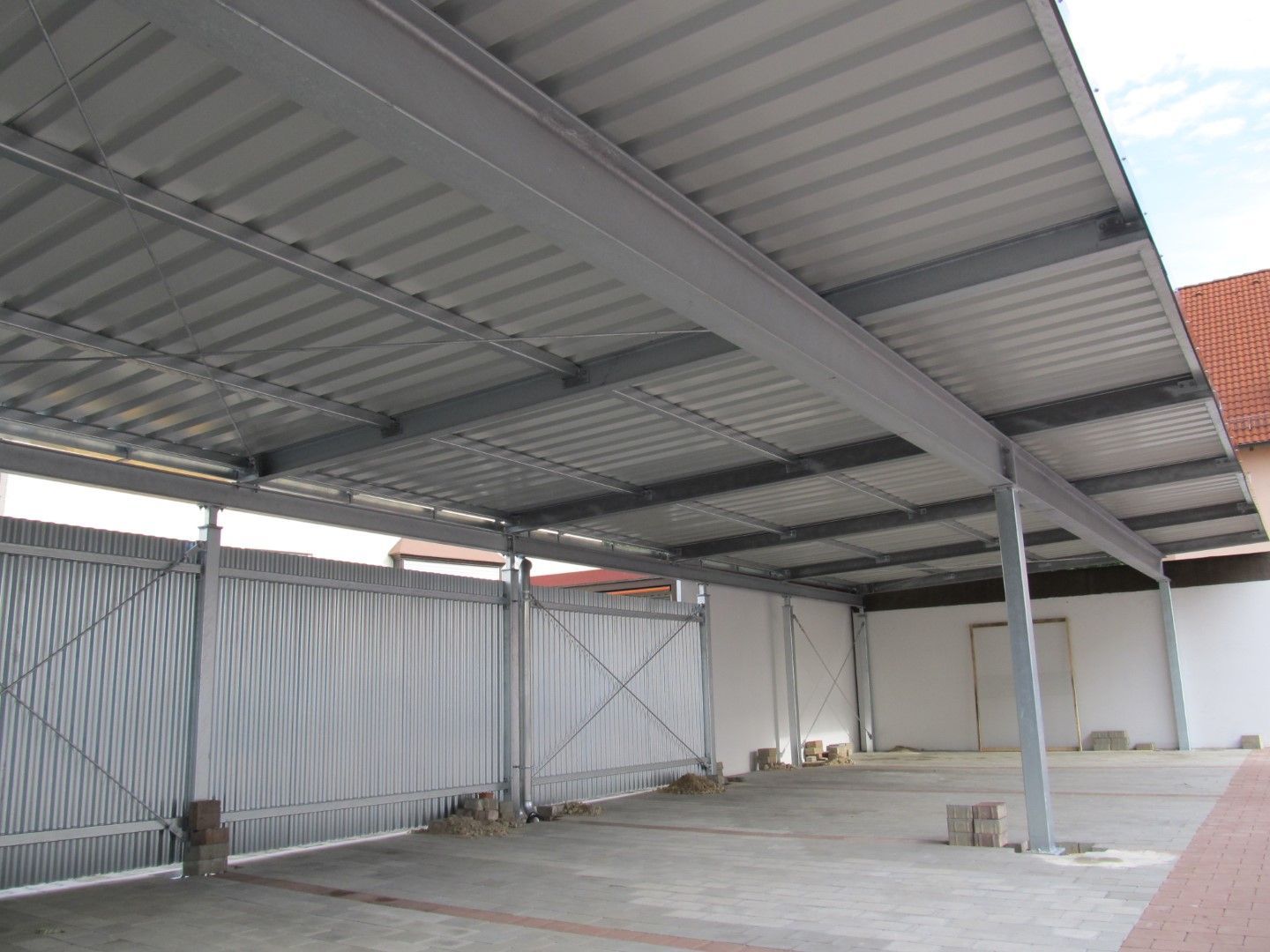Metallcarport mit Wellblechdach. Grauer Rahmen, Betonboden und teilweiser Metallzaun.