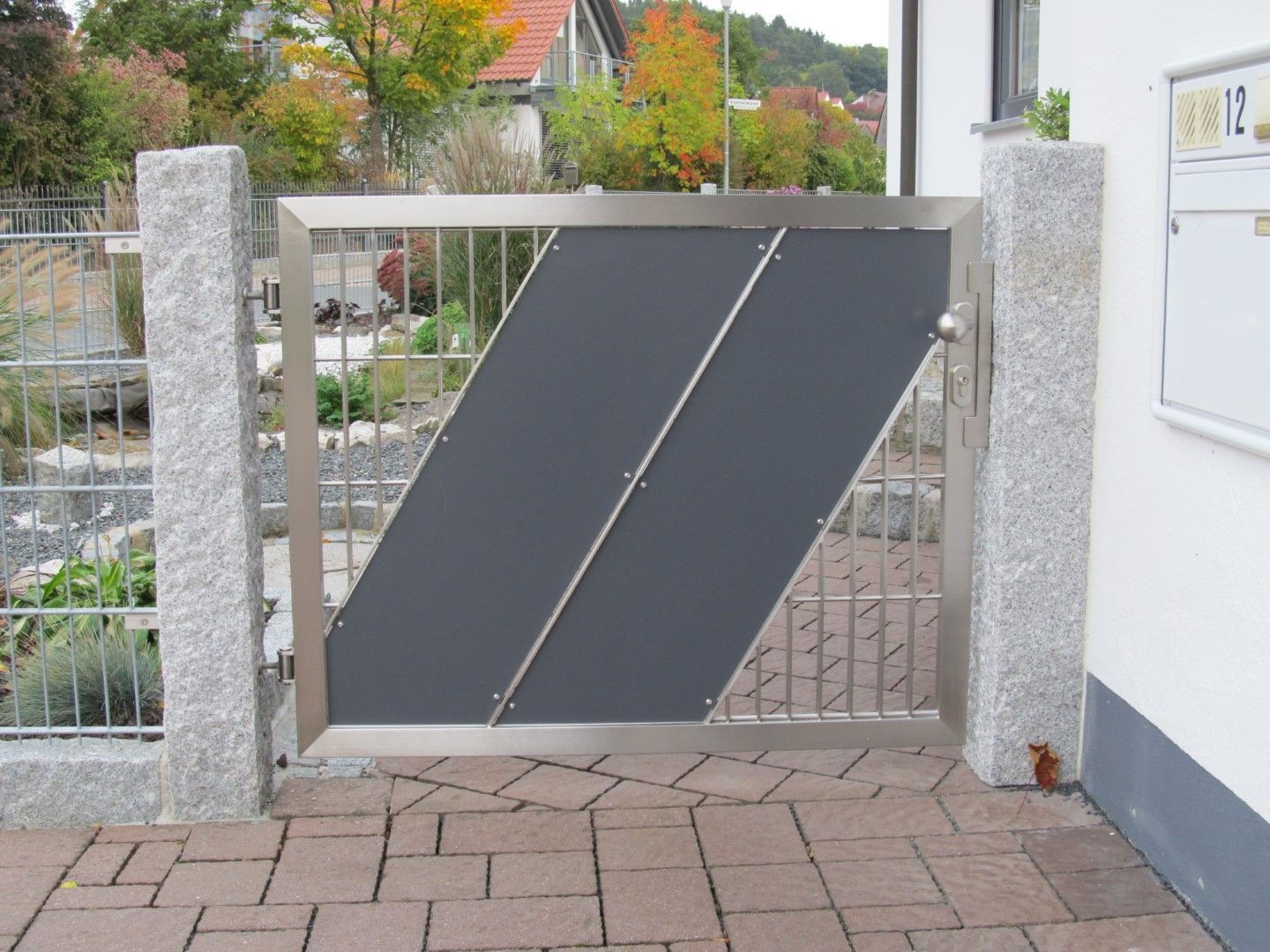 Ein modernes graues Metalltor mit diagonalen Paneelen zwischen Steinsäulen auf einem Ziegelweg.