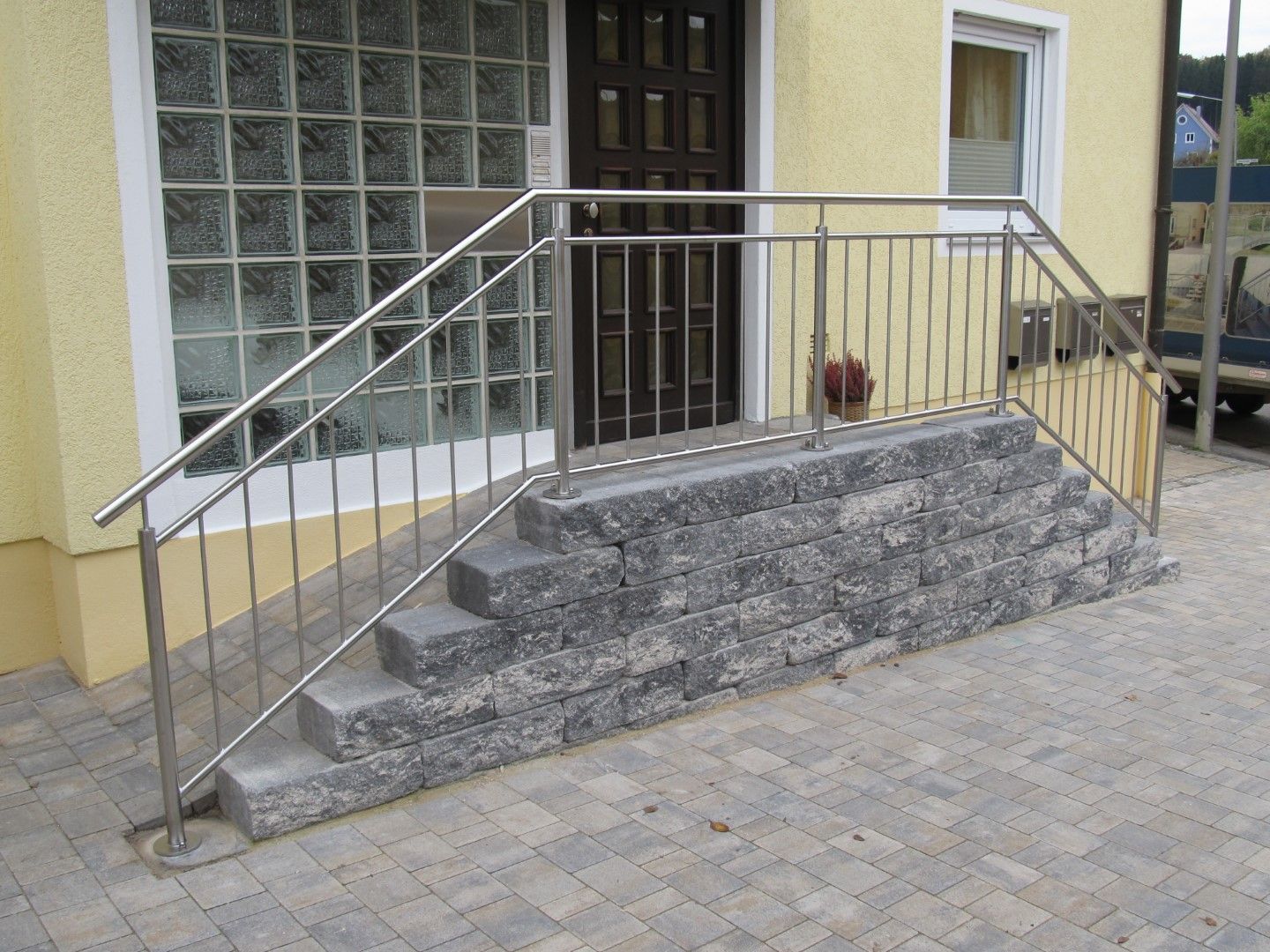 Handlauf aus Edelstahl neben Steinstufen, die zu einer braunen Tür führen. Gelbes Gebäude mit einem Glasbausteinfenster.