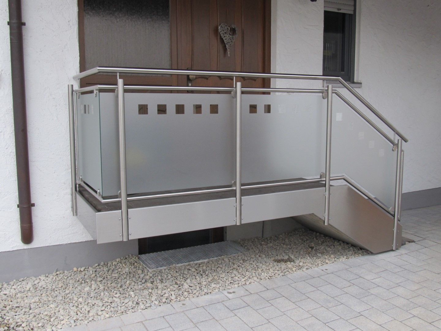 Rampe und Geländer aus Edelstahl mit Milchglas führen zu einer braunen Tür.