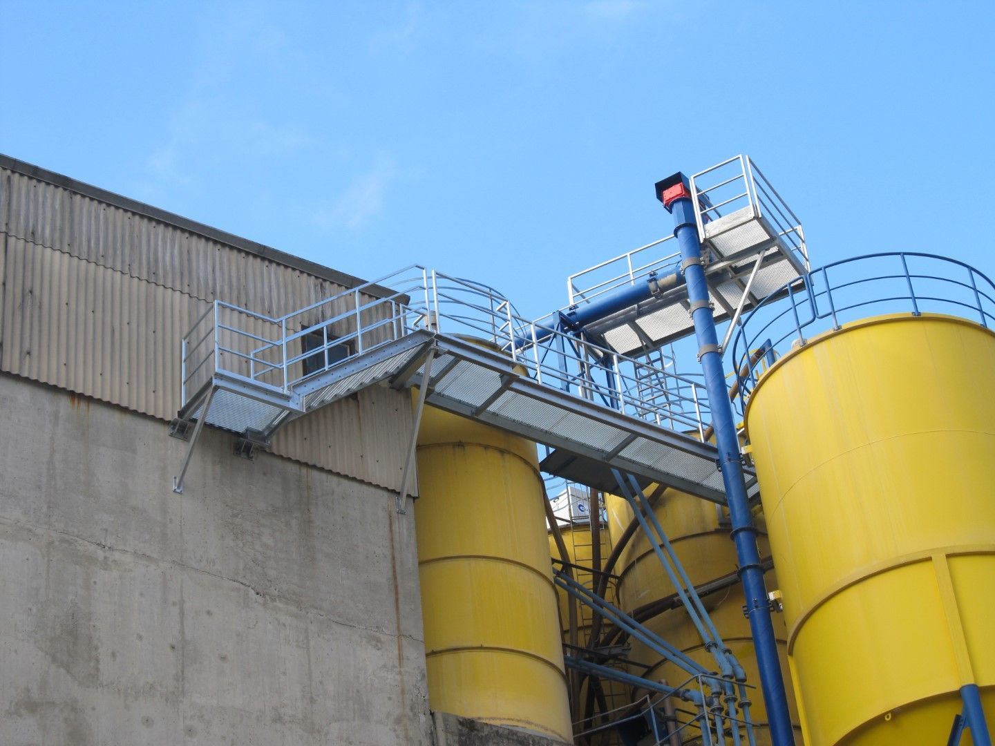 Industrieanlage mit gelben Tanks, blauem Förderband und erhöhtem Laufsteg vor blauem Himmel.