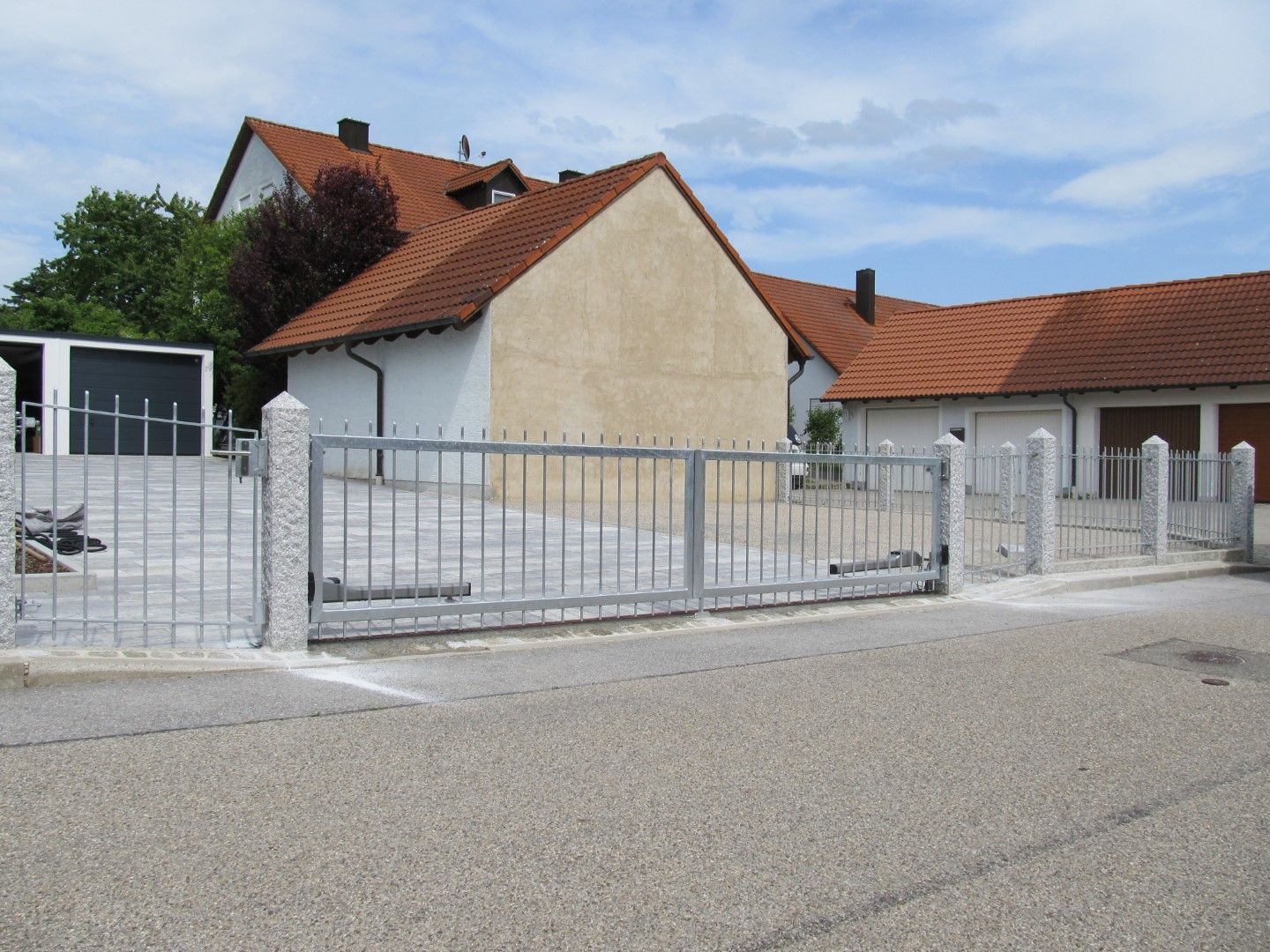 Graues Einfahrtstor aus Metall vor einem beigen Haus mit rotem Ziegeldach.