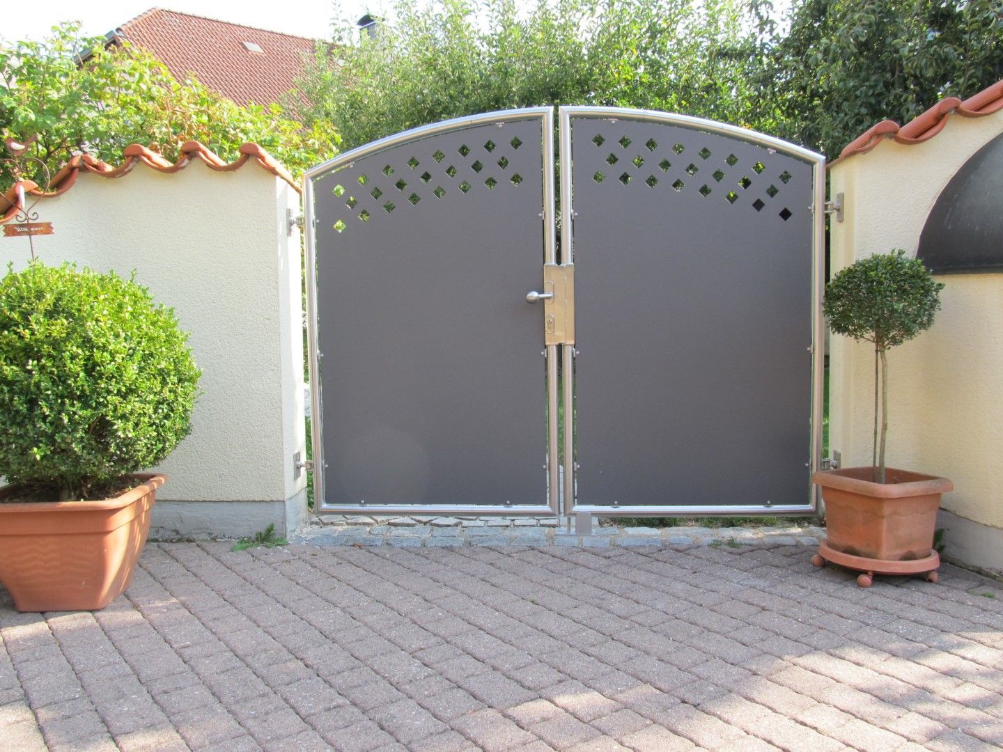 Graues Doppeltor mit Rautenausschnitten, flankiert von Topfsträuchern, in einer mit Ziegeln gepflasterten Auffahrt.