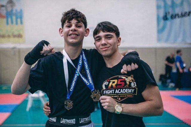 Dos luchadores con uniformes negros posan con sus medallas sobre una colchoneta en una academia de lucha.