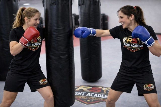 Dos mujeres con guantes de boxeo entrenando junto a sacos de boxeo en un gimnasio, sonriendo y dándose puñetazos.