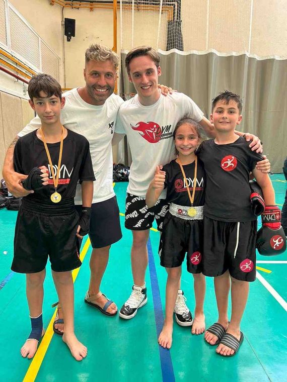 Cuatro estudiantes de artes marciales y dos adultos posando sobre una colchoneta azul de gimnasia con medallas y guantes
