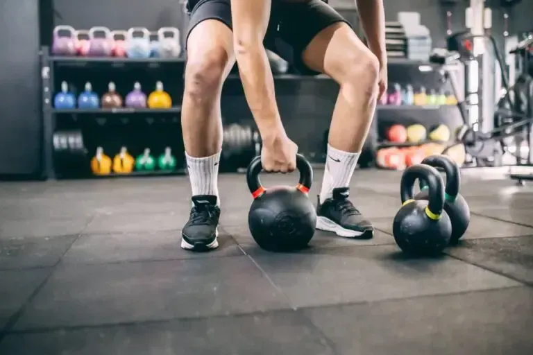 Una persona levanta una pesa rusa negra en un gimnasio con pesas rusas y mancuernas al fondo.