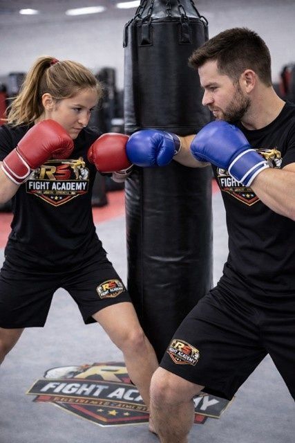Dos boxeadores con guantes rojos y azules entrenando alrededor de un saco de boxeo negro en un gimnasio.