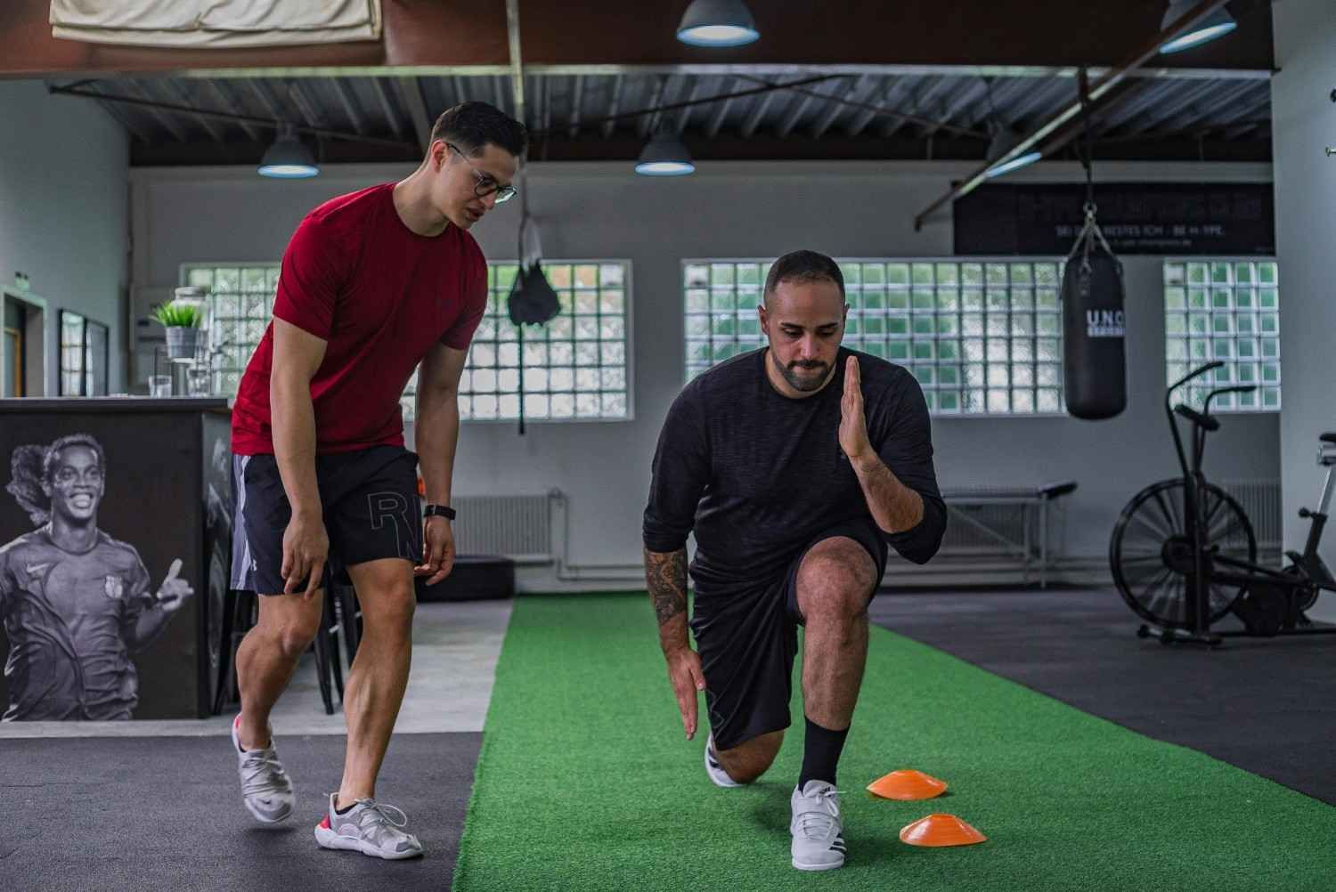 Dos hombres realizando zancadas de equilibrio en un gimnasio, con conos naranjas sobre una franja de césped artificial.