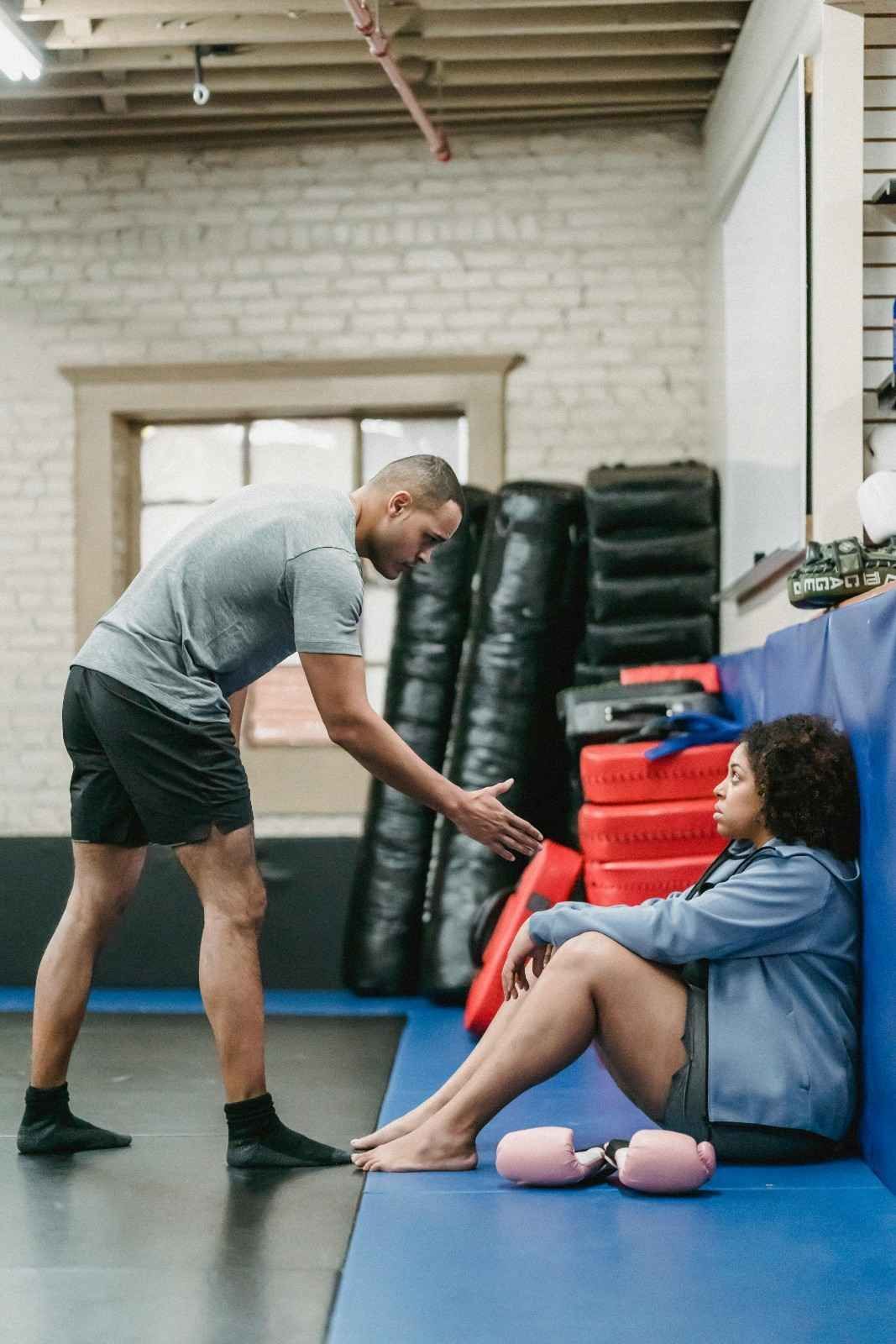 Un hombre ayuda a una persona sentada a estirar las piernas en un gimnasio o espacio de entrenamiento.
