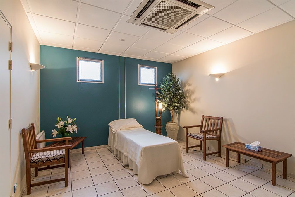 Pièce funéraire avec un cercueil, des chaises et une plante. Mur turquoise et sol carrelé blanc.