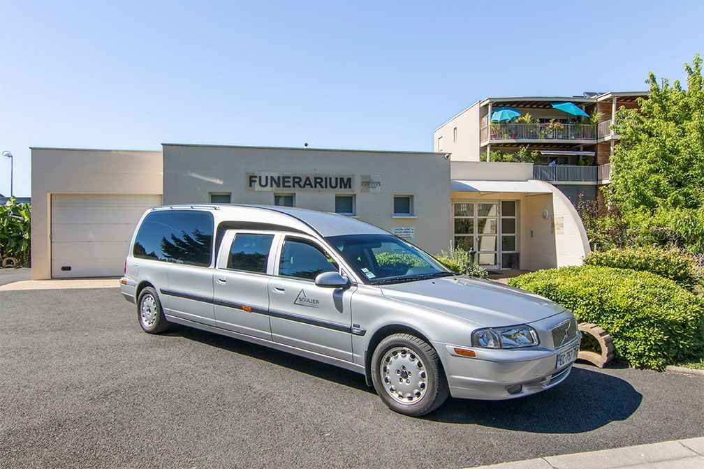 Un corbillard argenté est stationné devant un bâtiment moderne portant l'inscription « Funérarium » par une journée ensoleillée.