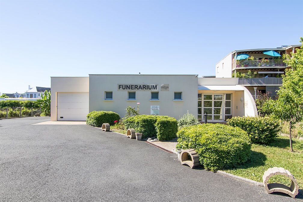 Extérieur d'une maison funéraire moderne avec une porte de garage, des buissons verts et l'inscription « FUNÉRAILLES » au-dessus de l'entrée.