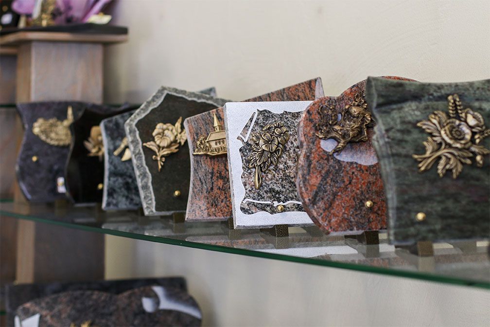 Pierres tombales sur une étagère en verre. Formes et couleurs variées, ornées de décorations métalliques.