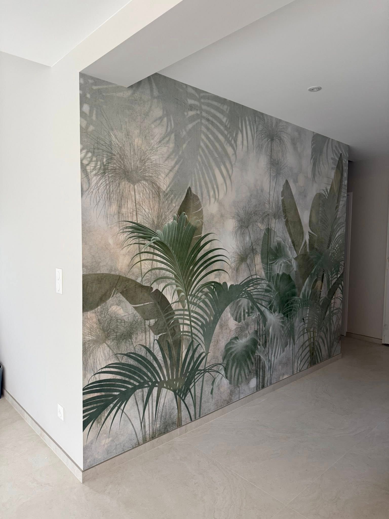 Un grand mur d'accent dans une pièce présente une fresque murale représentant de luxuriantes feuilles de palmier tropicales vertes sur un fond gris doux et brumeux.
