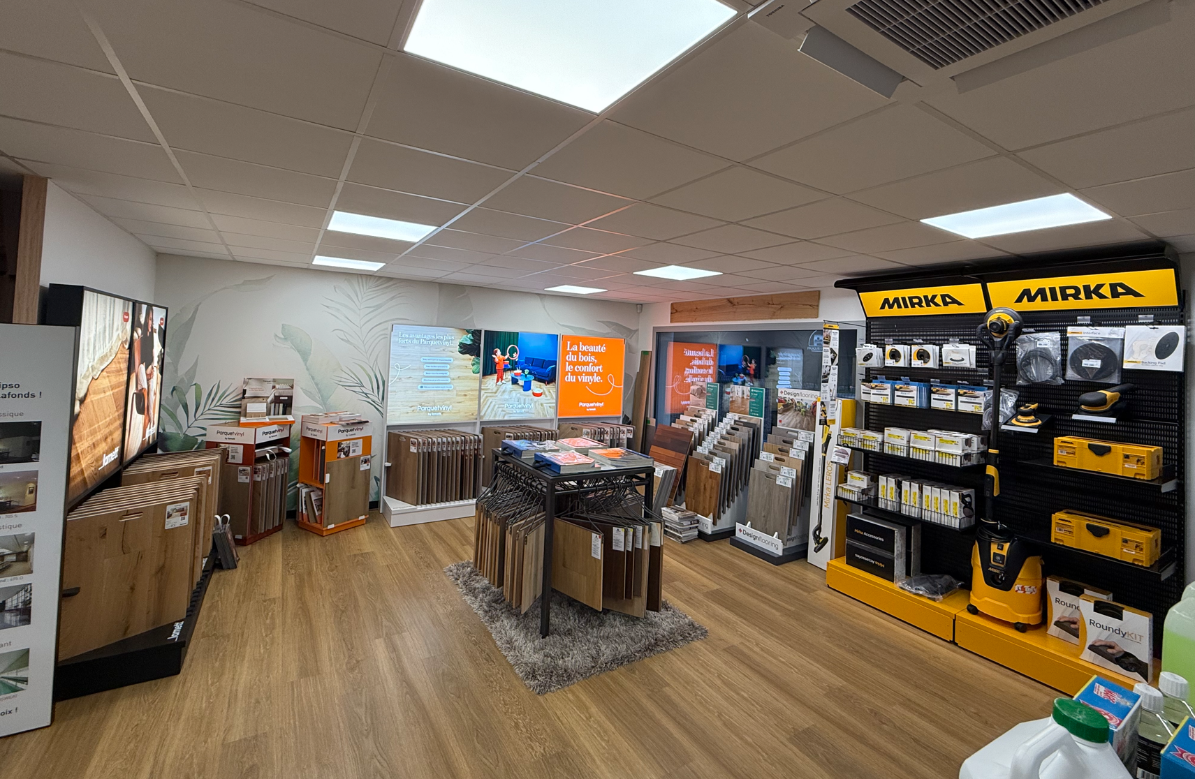 Intérieur d'un magasin de bricolage ou de revêtements de sol, avec des présentoirs de bois et divers outils électriques jaunes.