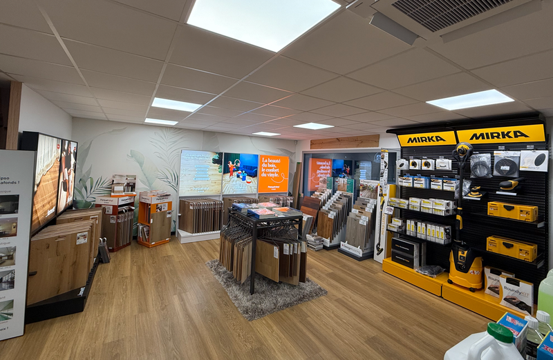 Un espace d'exposition présentant des échantillons de revêtements de sol, des présentoirs de produits et des étagères jaunes remplies d'équipements.