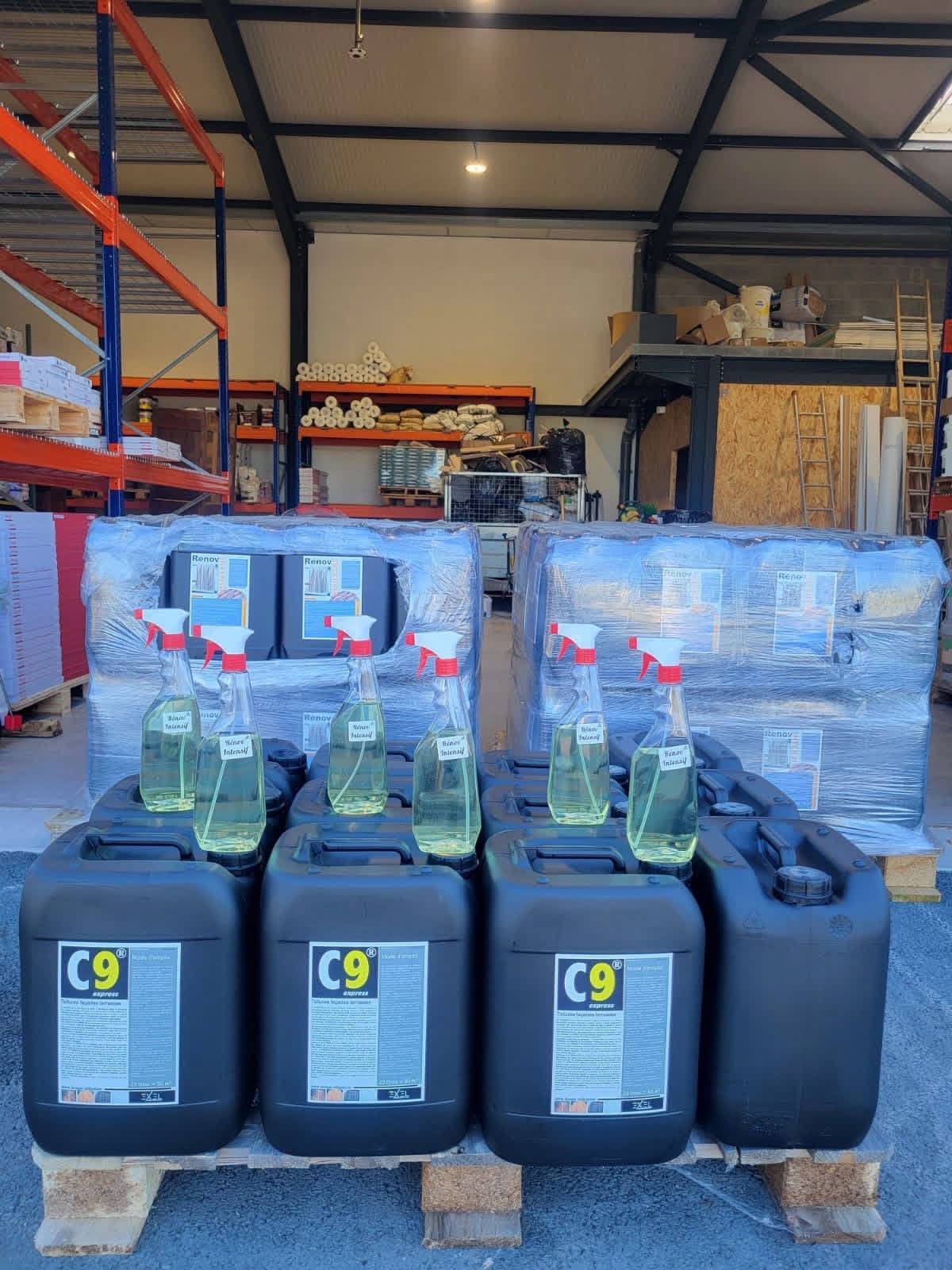 Quatre bidons de nettoyage C9 en plastique noir, munis de flacons pulvérisateurs, sont posés sur une palette d'expédition dans une zone de stockage d'un entrepôt.