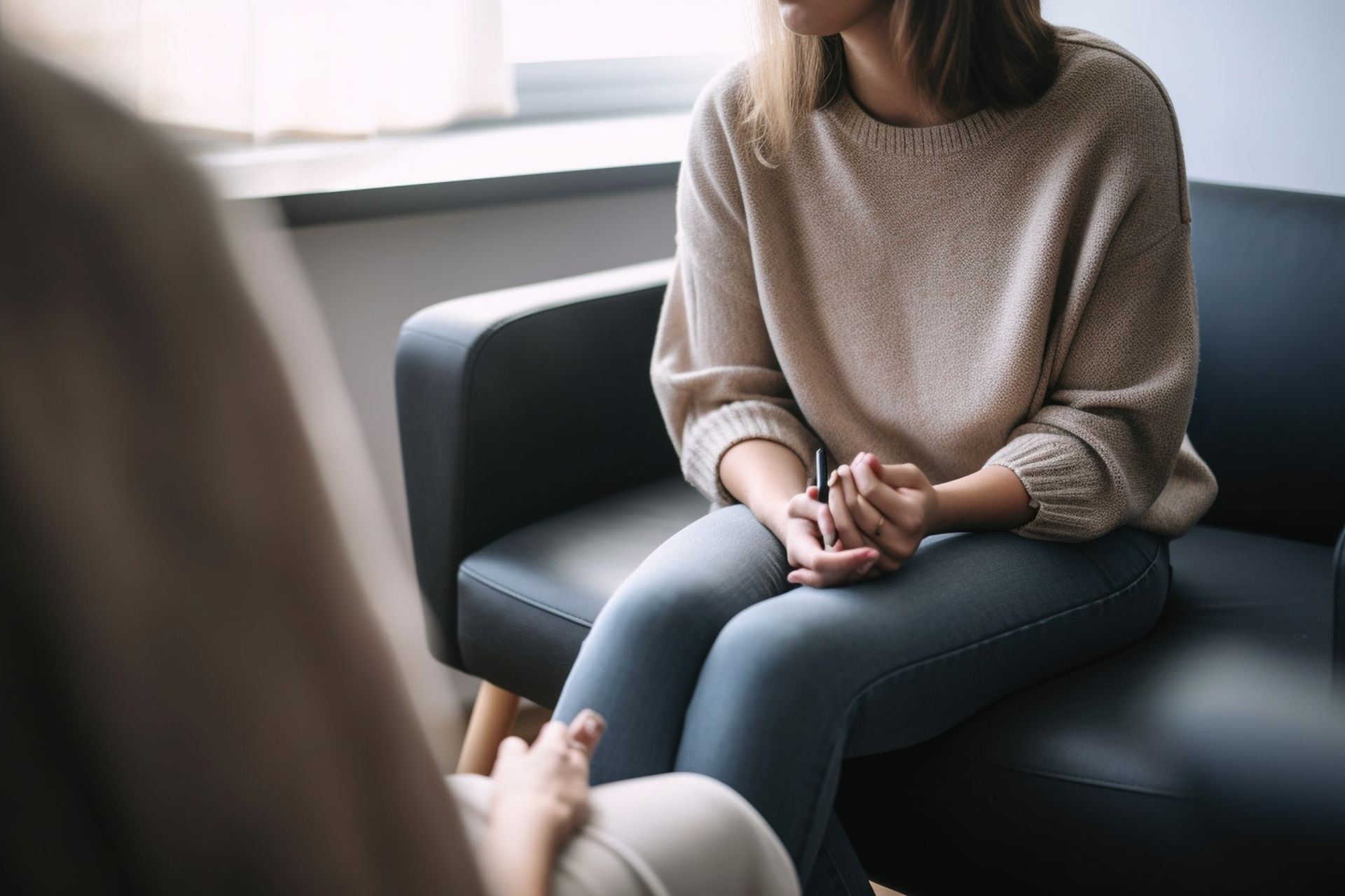 Femme assise sur un canapé, face à une autre personne, les mains jointes, à l'intérieur d'une pièce.