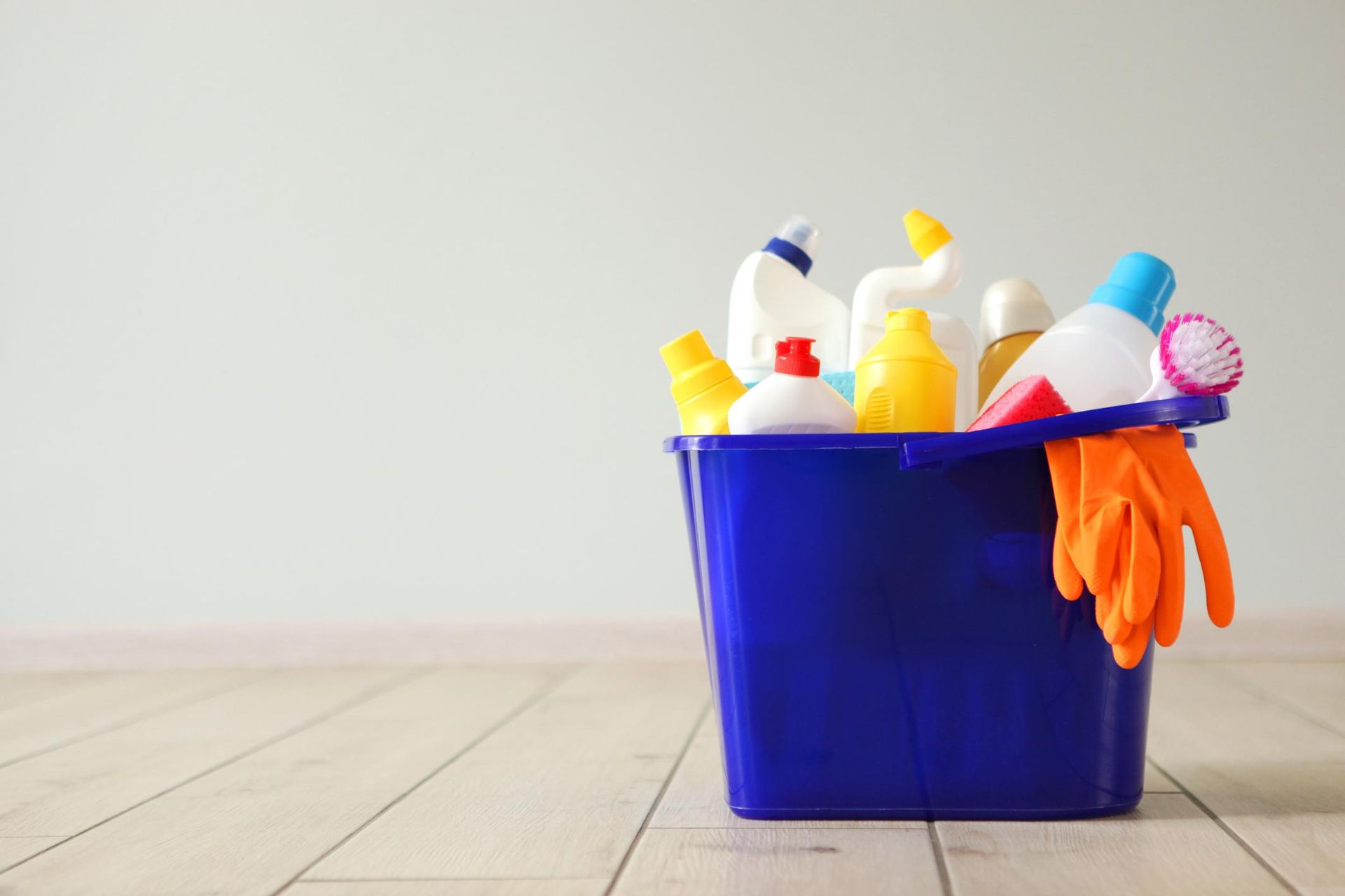 Cesta de limpieza azul con botellas pulverizadoras, esponjas y guantes de goma naranjas sobre un suelo de madera.