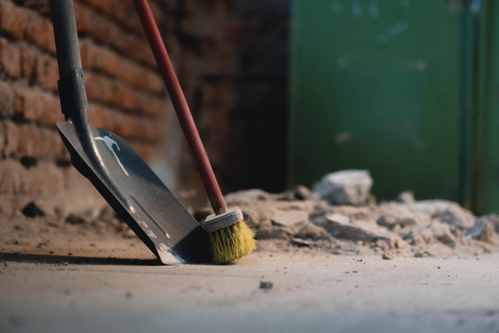 Escoba y recogedor sobre un suelo polvoriento junto a una pared de ladrillos y un contenedor verde.