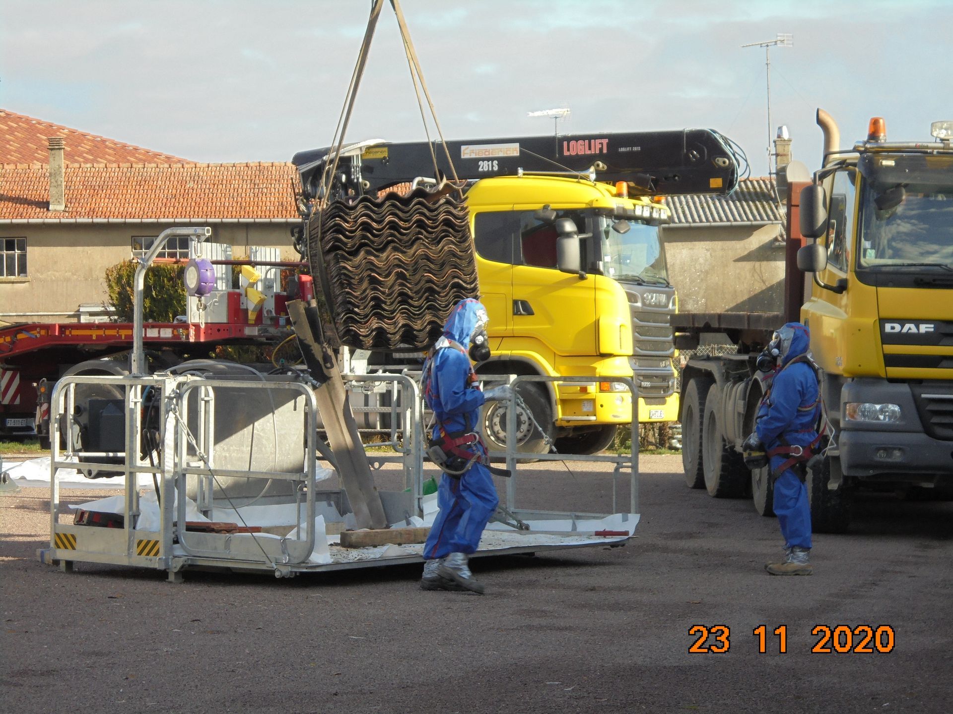 Grue en train de soulever un tas d'ancienne tuile en amiante avec deux personnes en bleues en combinaison devant