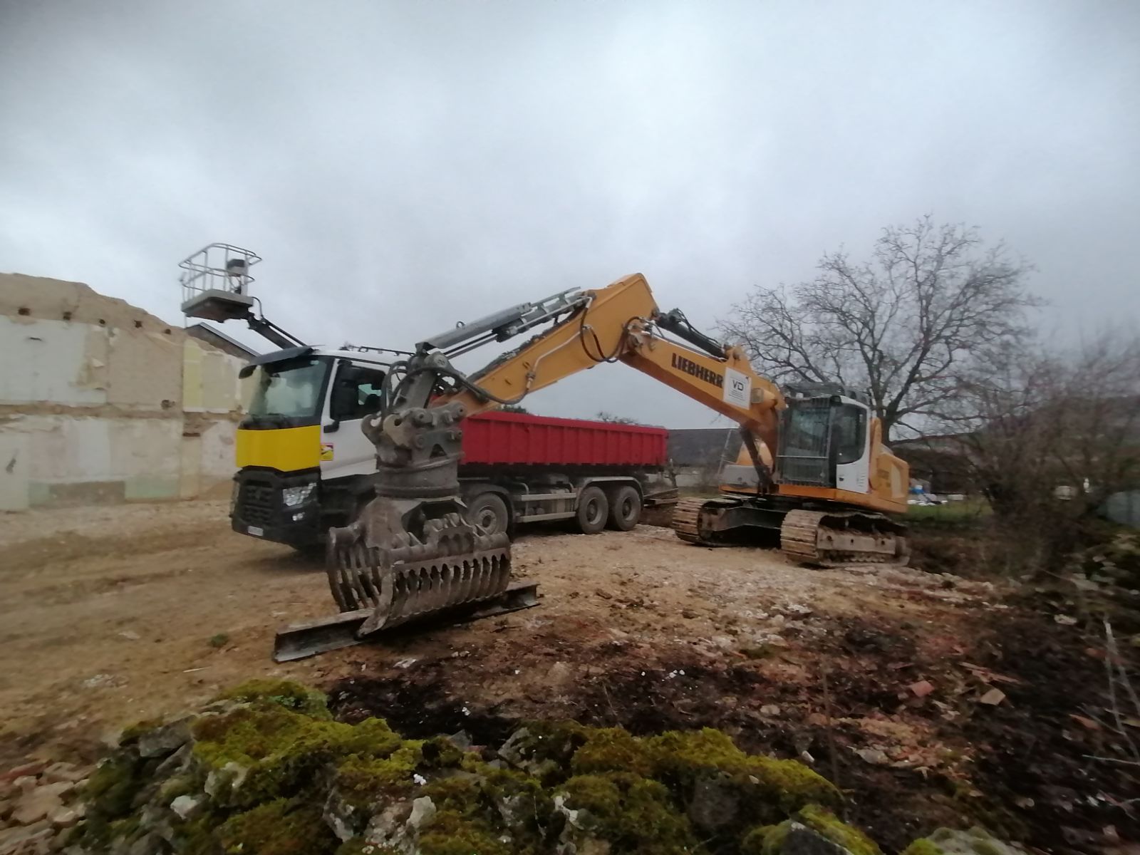 Tractopelle Liebherr jaune avec pince en train de prendre une barre de métal et camion avec benne rouge en arrière-plan