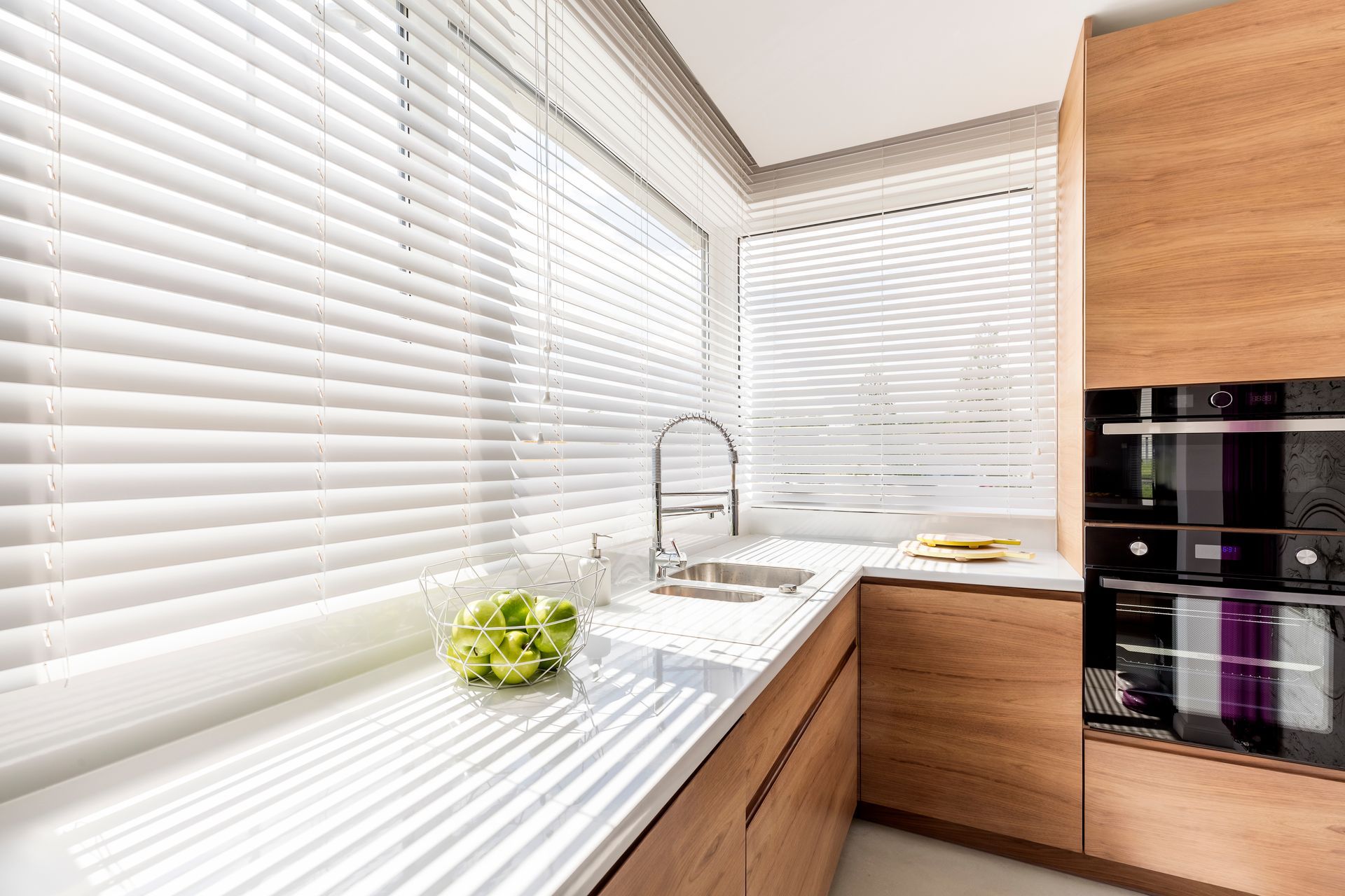 Cuisine moderne et lumineuse avec stores blancs, placards en bois, évier en inox et four encastré.