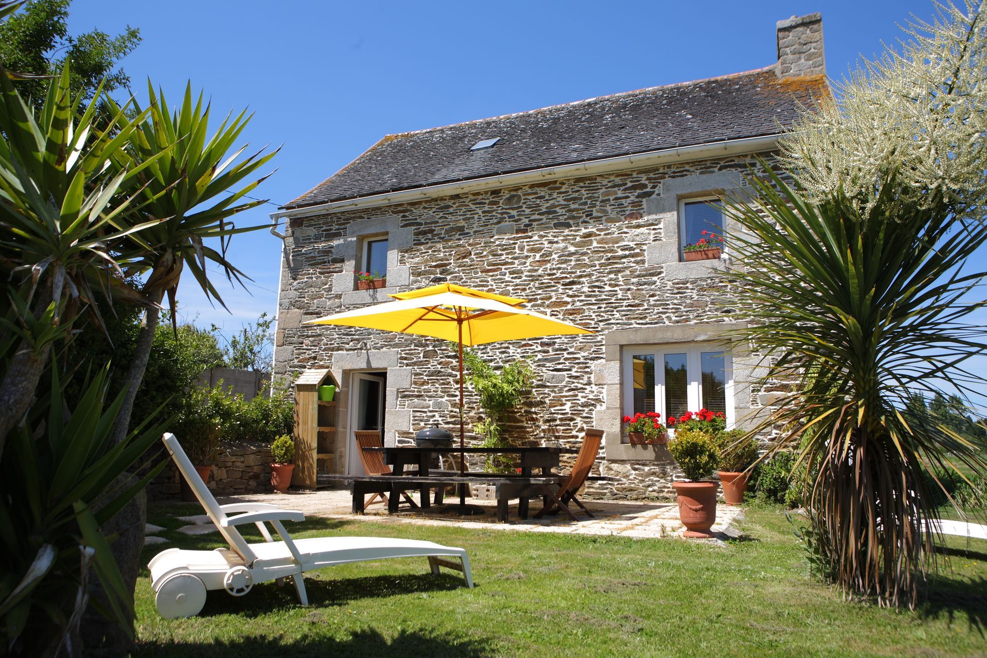 Jardin de cottage en pierre avec table de patio, parasol jaune et chaises longues au soleil