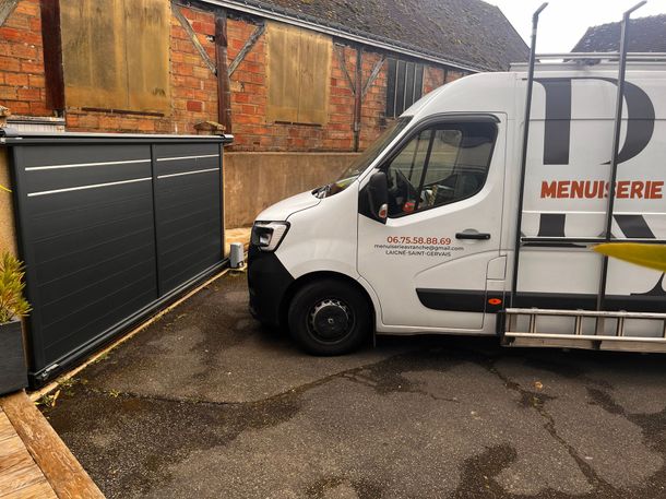 Une camionnette de Menuiserie Avranche garée à côté d'un portail noir.