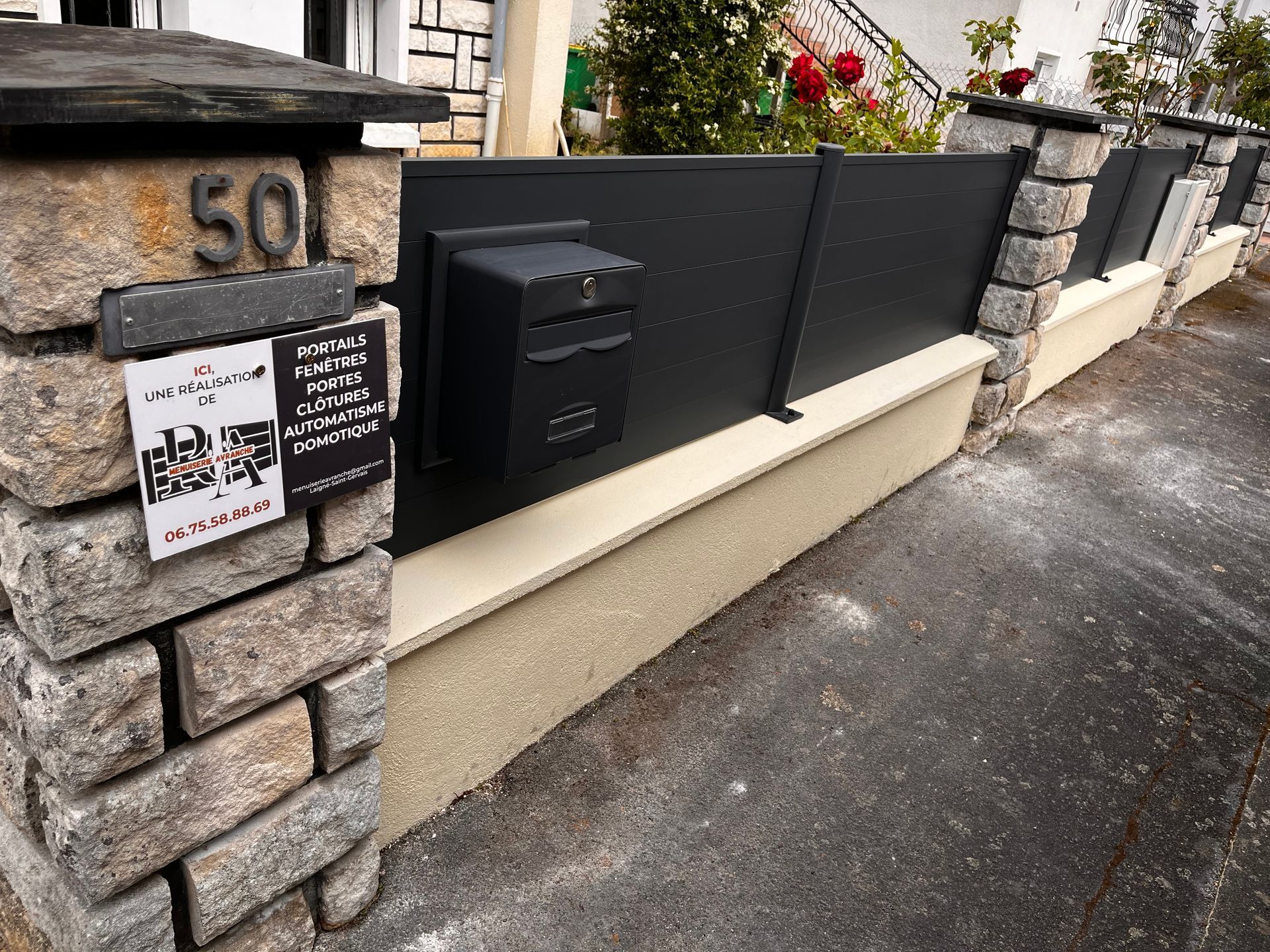 Boîte aux lettres noire et portail sur un mur en pierre à côté d'une maison.