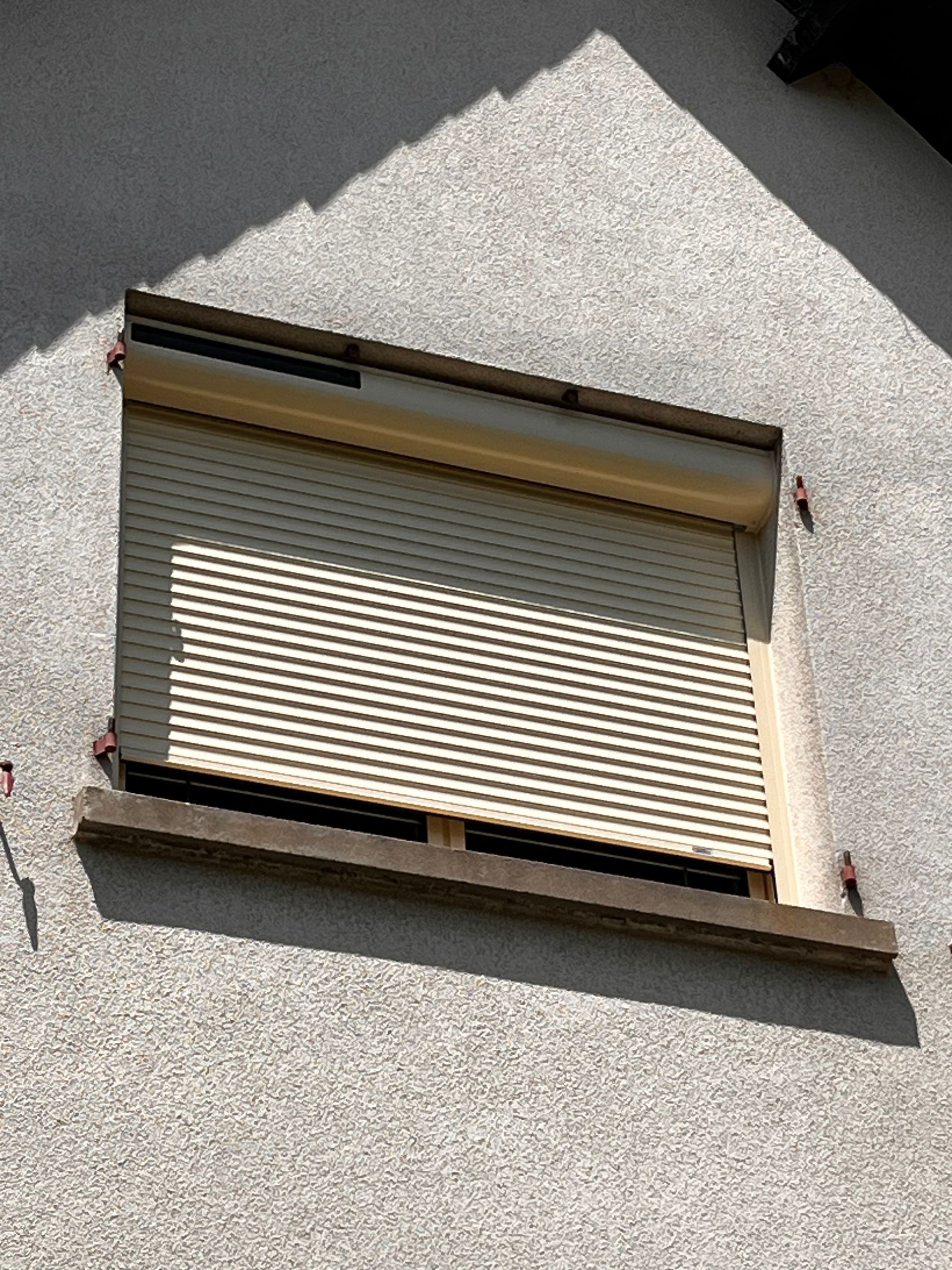 Volet beige fermé sur un mur en stuc texturé, baigné de soleil.
