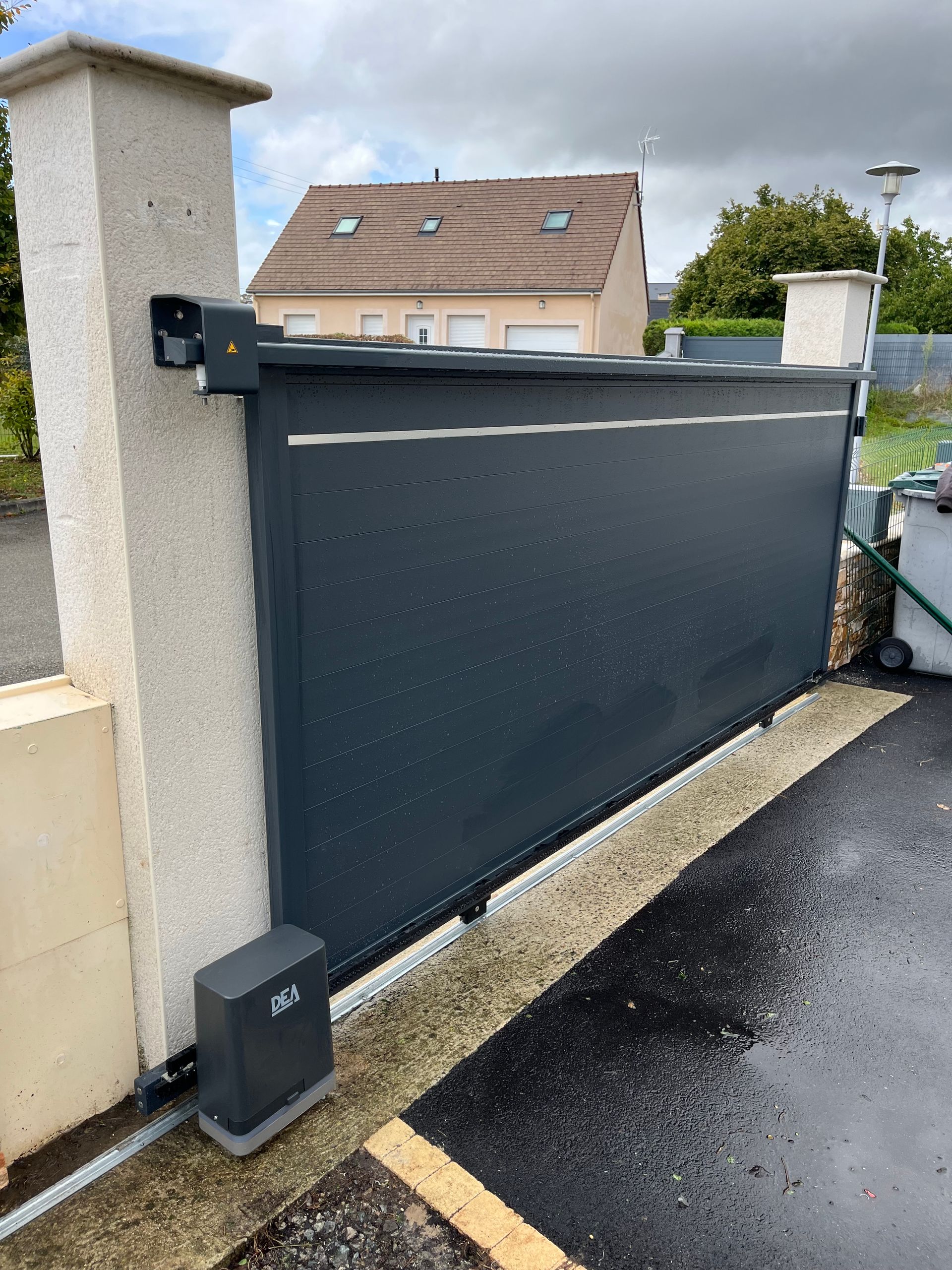 Portail de garage rétractable noir à côté d'un pilier blanc et d'asphalte mouillé dans une cour résidentielle.