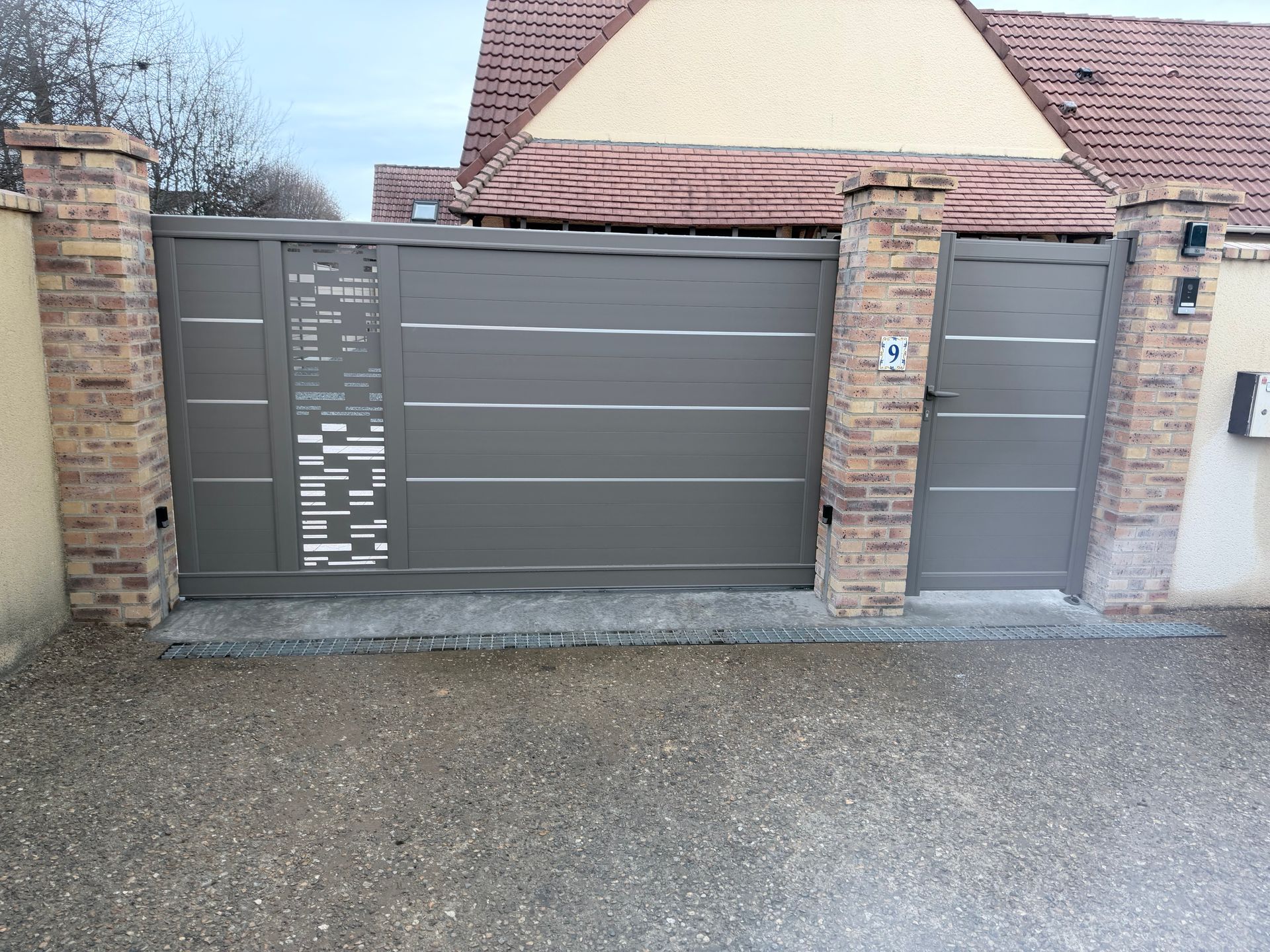 Portail d'allée et porte piétonne modernes en métal gris, encastrés dans un mur de briques à côté d'une allée de maison.