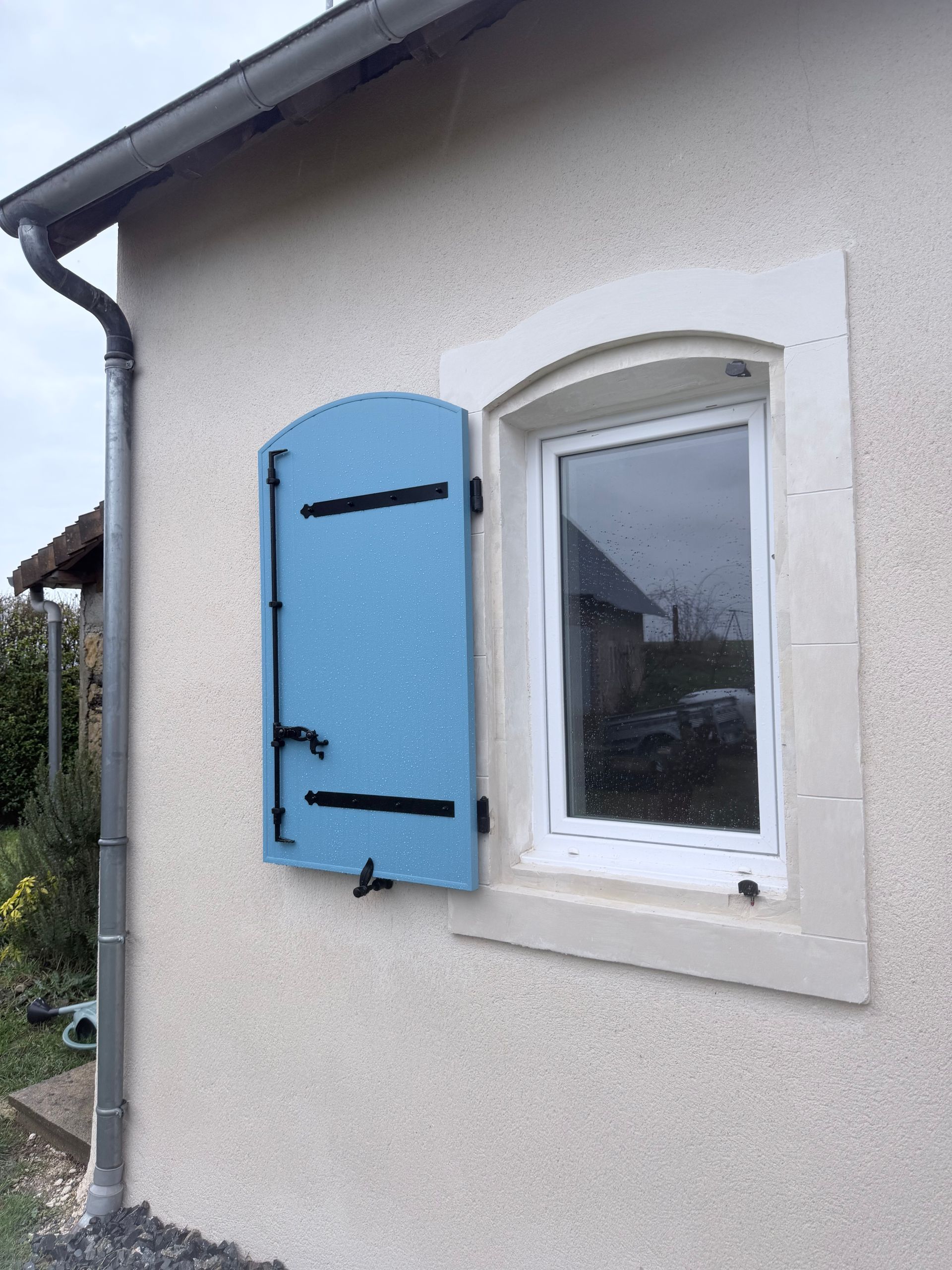 Un volet bleu sur une maison au crépi blanc.