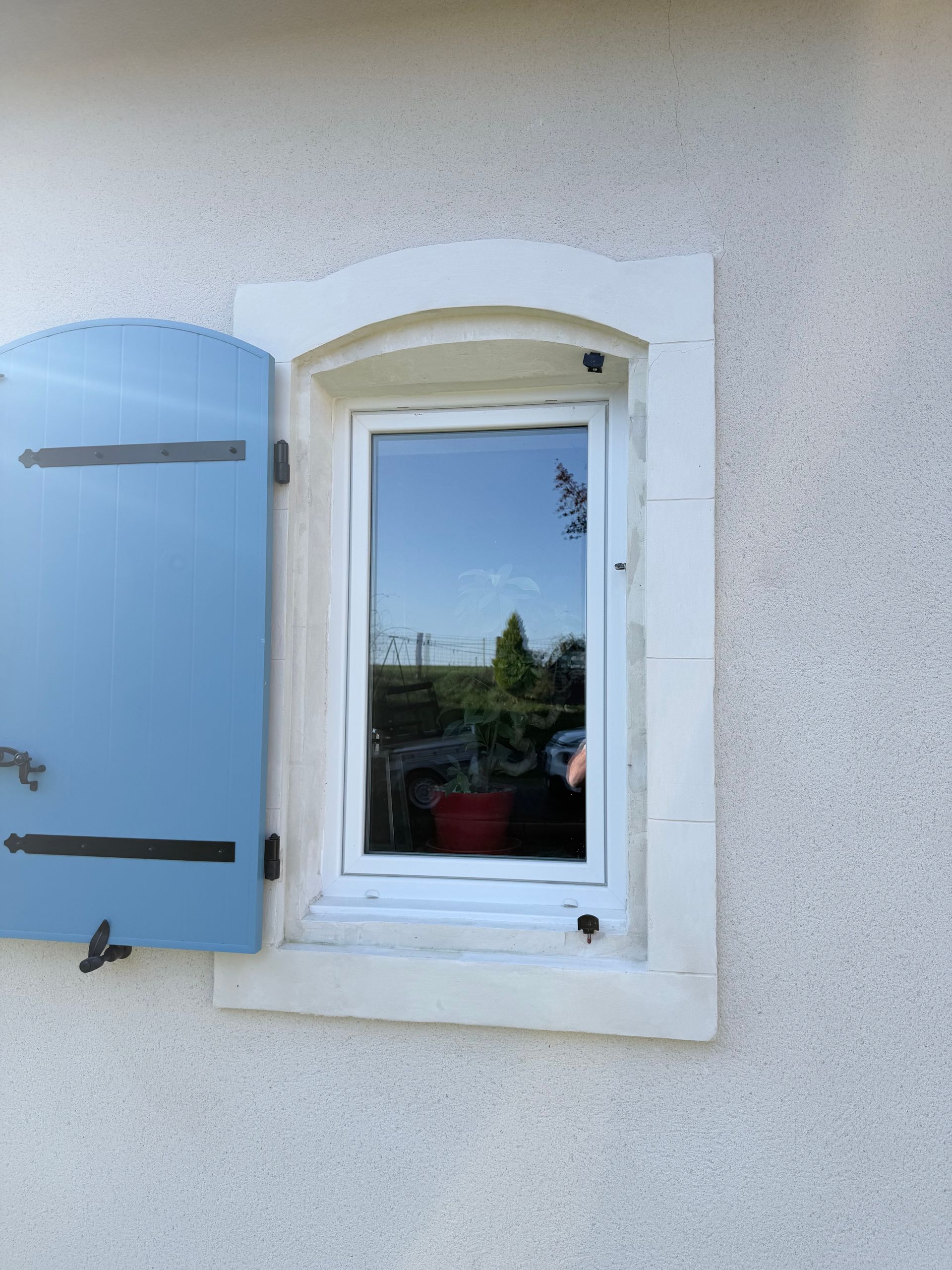 Fenêtre blanche à volet bleu sur un mur en stuc, reflétant les arbres et le ciel.