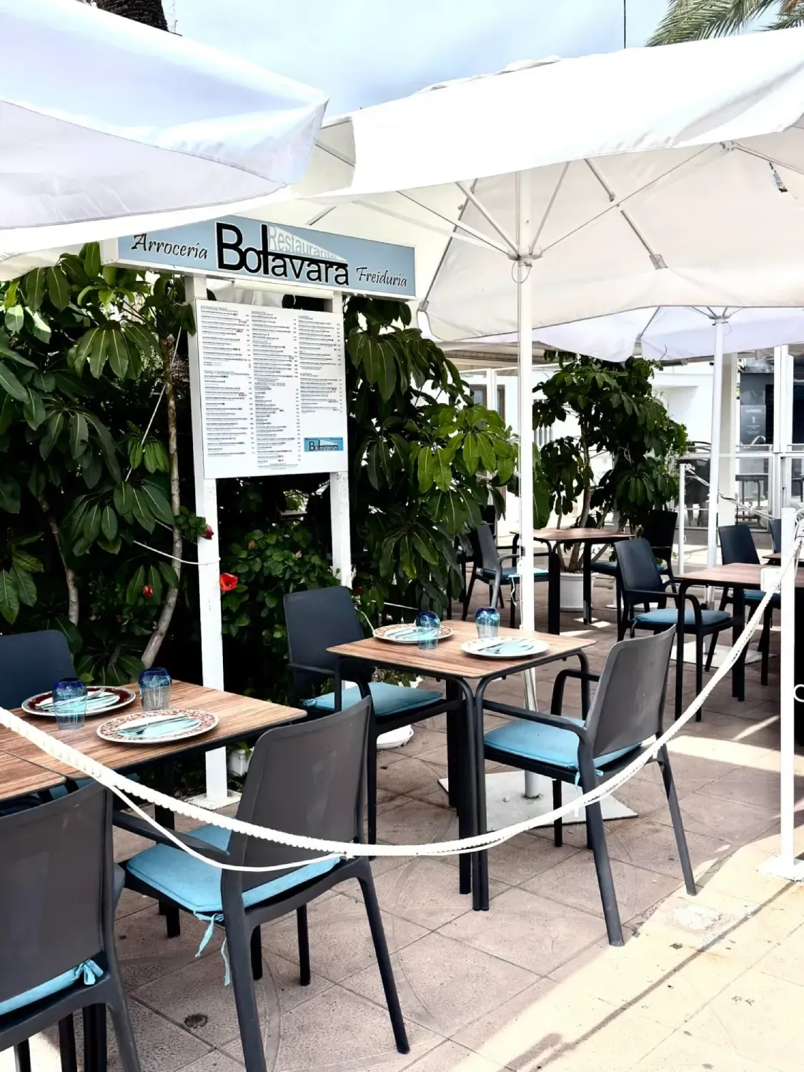 Terraza de cafetería con mesas pequeñas, sillas negras y una pizarra con el menú.