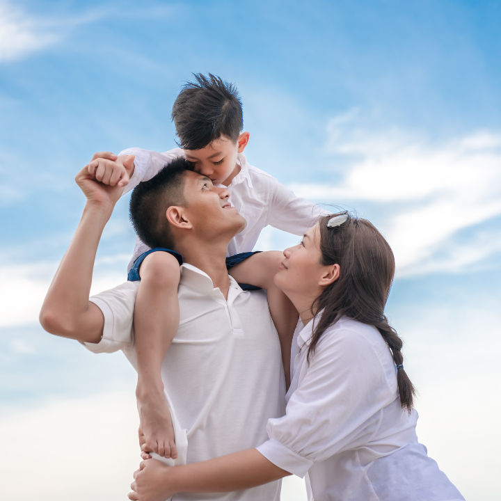 Familia de tres bajo un cielo azul, el hijo sobre los hombros del padre besándole la cabeza, la madre abrazando al padre.