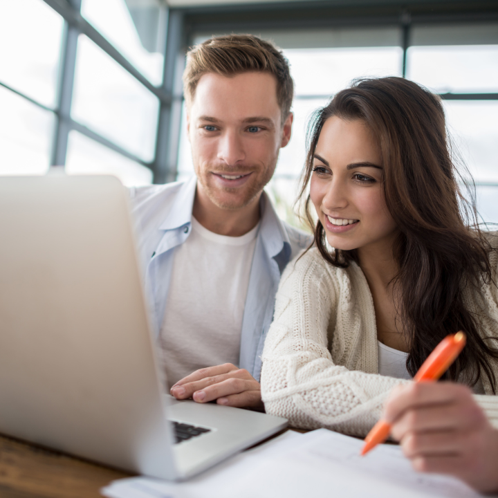 Una pareja sonriendo mientras usa una laptop. Una mujer escribe con un bolígrafo naranja, con papeles sobre el escritorio.
