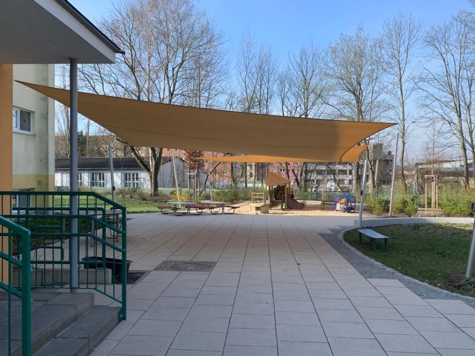 Außenbereich mit Sonnensegel über dem Spielplatz. Hellbraunes Sonnensegel, gefliester Boden, Bänke und Bäume.