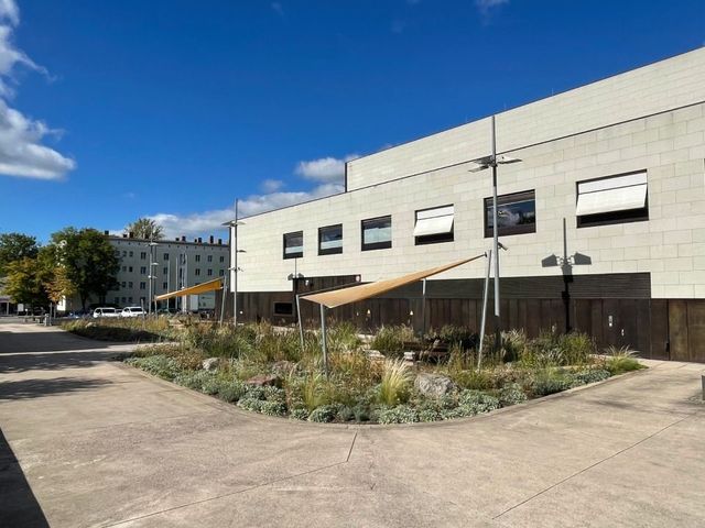 Außenansicht eines modernen Gebäudes mit Garten. Das Gebäude ist hellfarbig mit dunklen Fenstern und Markisen.