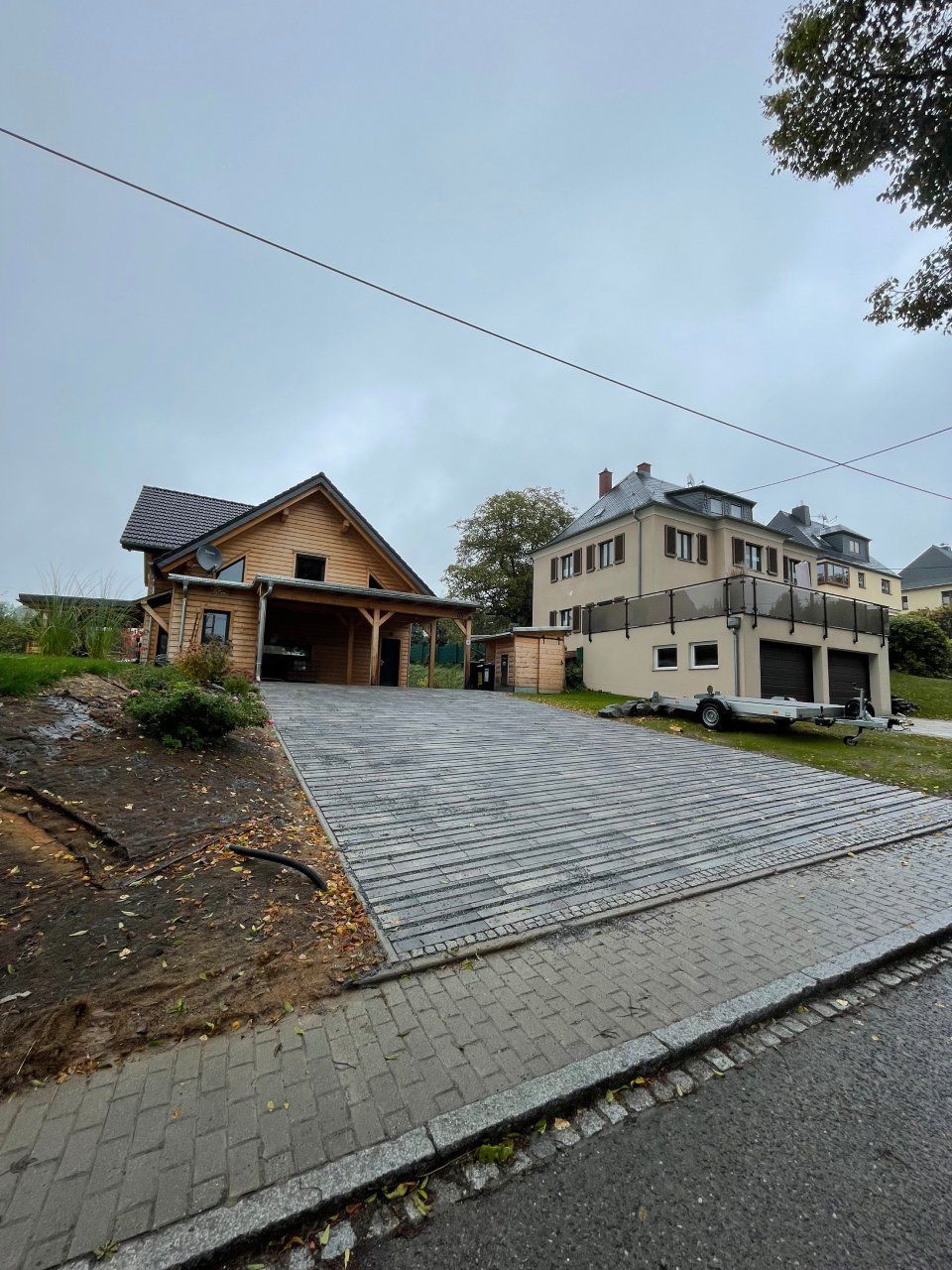 Zwei Häuser mit gepflasterten Auffahrten an einem bewölkten Tag. Das linke Haus ist aus Holz und hat einen Carport. Das rechte ist verputzt.