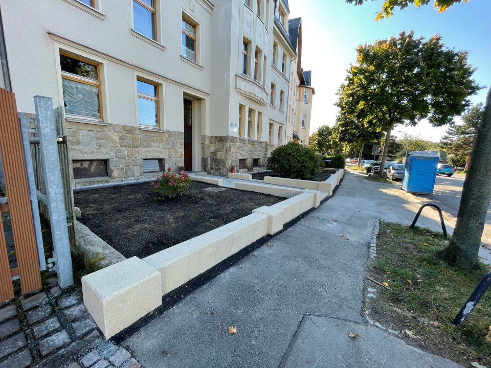 Blick vom Bürgersteig auf das Gebäude mit niedrigen Betonpflanzkästen und kleinem Garten. Eine blaue mobile Toilette ist zu sehen.