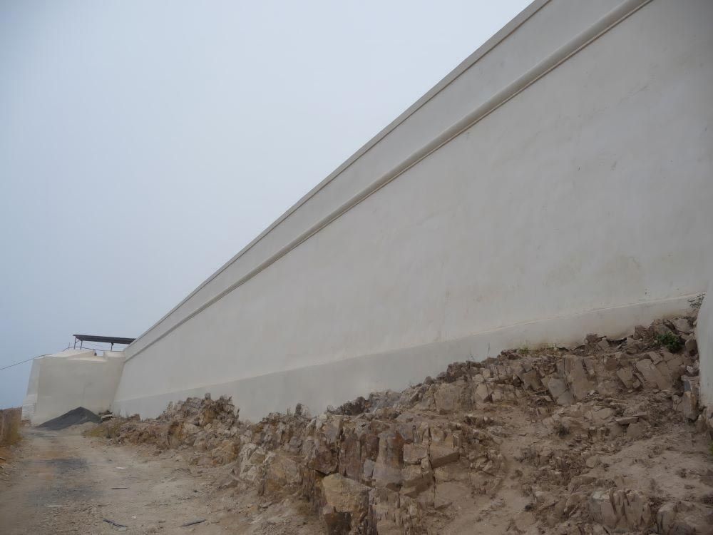 Largo muro de contención blanco a lo largo de un terraplén de tierra fangosa bajo un cielo gris.