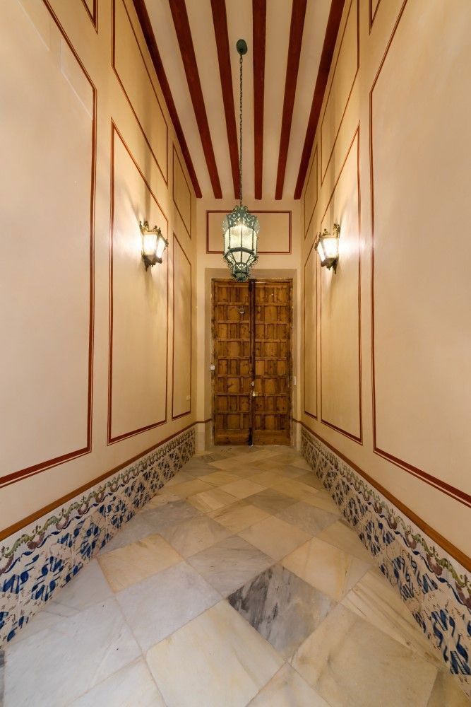 Estrecho pasillo alicatado con paredes color crema, molduras azules, puerta de madera y farol colgante.