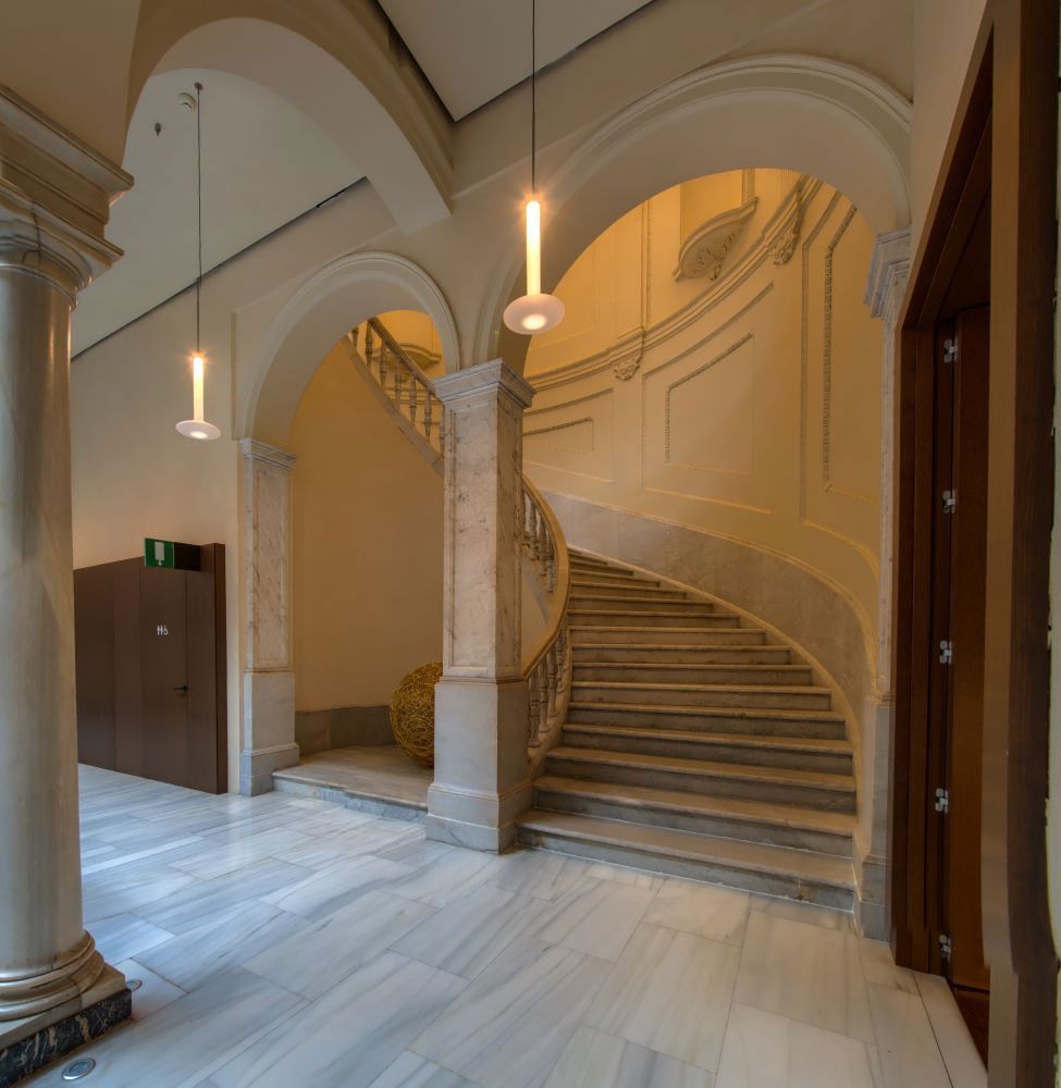 Elegante escalera interior en un pasillo abovedado con techos arqueados, columnas de piedra y lámparas colgantes.