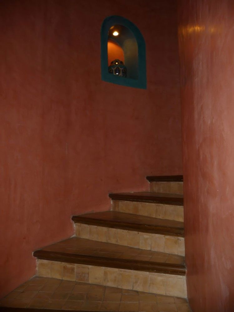 Escalera curva en un estrecho pasillo de terracota, con una pequeña lámpara de techo azul en la pared.