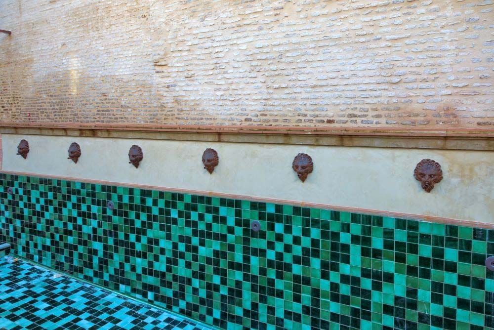 Pared de la piscina con mosaico de color turquesa y piedra blanca texturizada en la parte superior, bordeada de pequeñas fuentes con forma de cabeza de león.
