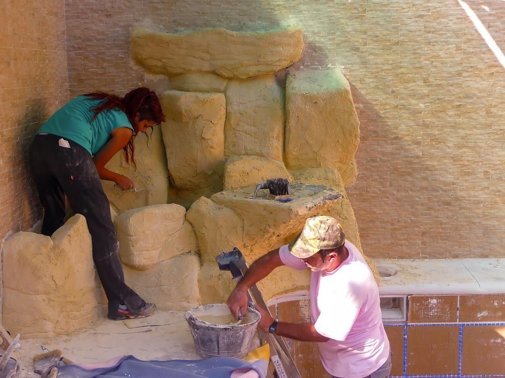 Dos obreros dando forma a un muro de piedra, uno subido a una escalera y el otro agachado con herramientas.