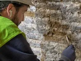 Un trabajador con un chaleco verde neón inspecciona un muro de piedra rugosa con una herramienta en el interior de un edificio.